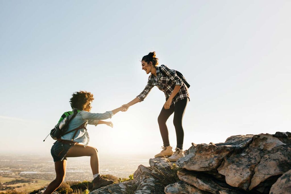a woman helps another woman up a rock on a hike