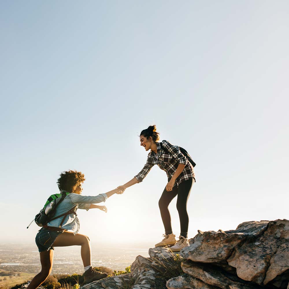 a woman helps another woman up a rock on a hike